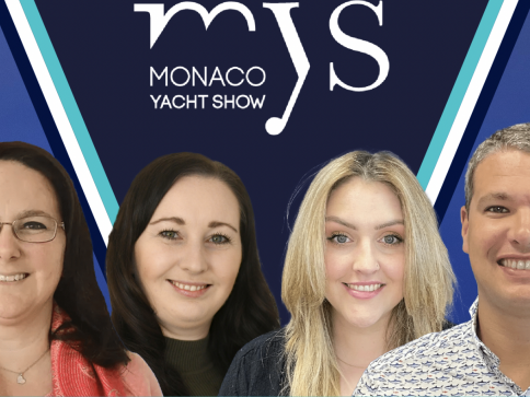 Four people, three women and one man, are smiling in front of a blue background featuring the Monaco Yacht Show (MYS) logo in white letters. The group is framed by a white and turquoise V-shaped design.