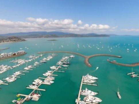 Aerial view of a marina with numerous white yachts moored in turquoise water, curved breakwaters, scattered boats on the sea, and distant mountains under a partly cloudy sky.