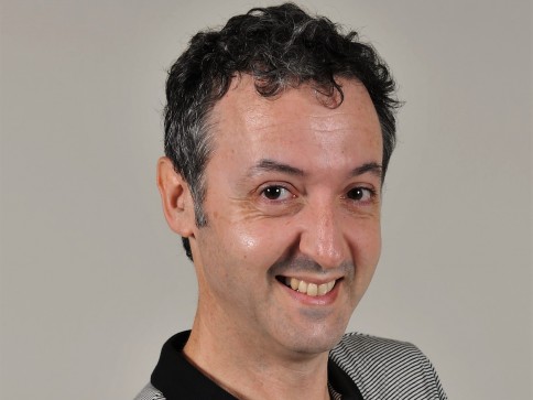 A man with short, curly dark hair and a friendly smile looks slightly to the side. He is wearing a collared shirt with black and white stripes and is set against a plain, light background.