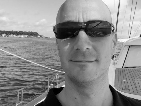 A man wearing sunglasses takes a selfie on a boat, with water and a distant shoreline visible in the background. The image is in black and white.