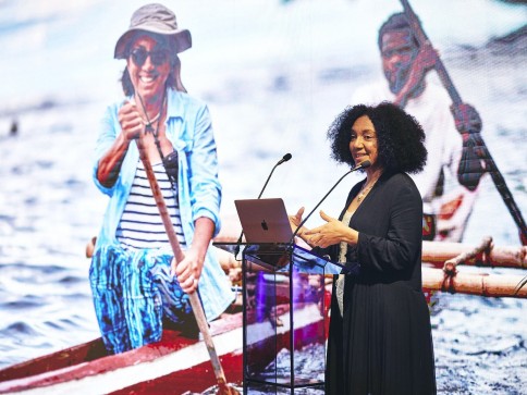 A woman stands at a lectern speaking into microphones, with a large screen behind her displaying an image of a smiling person paddling a canoe on the water.