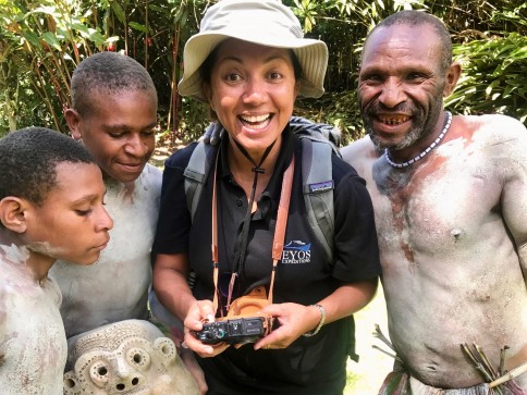 A smiling woman wearing a hat and rucksack holds a camera whilst standing with three men in traditional attire and body paint, surrounded by lush greenery.
