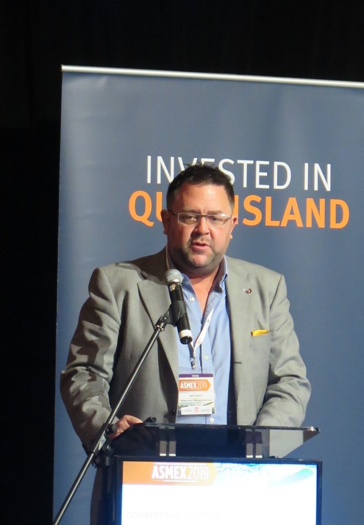 A man in a suit speaks at a lectern with a microphone. Behind him is a banner that reads INVESTED IN QUEENSLAND. He wears glasses and has conference badges around his neck.