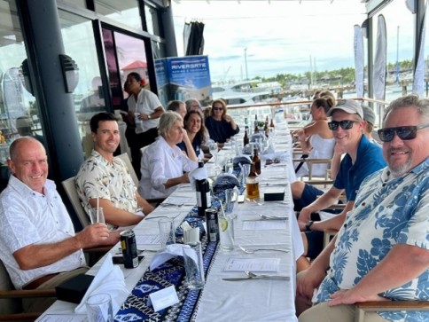 A group of people sit around a long table set for a meal at an outdoor restaurant by a marina, smiling at the camera. The background shows boats and palm trees on a sunny day.