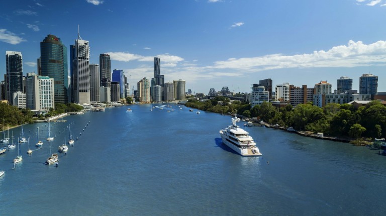 A wide river flows through a city with modern high-rise buildings on the left, trees on the right, and several boats, including a large white yacht, on the water under a blue sky with scattered clouds.