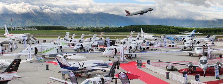 A busy airport apron displays several private jets and small aeroplanes parked side by side, with a commercial plane taking off in the background under a cloudy sky and mountains.