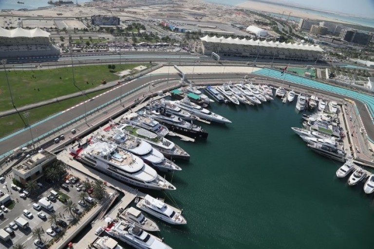 Aerial view of a marina filled with yachts beside a race track, with green spaces, parked cars, buildings, and the sea visible in the background.