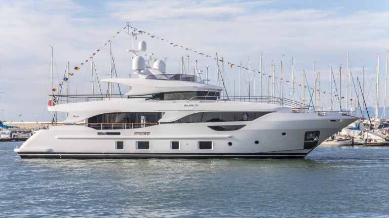 A large white luxury yacht with multiple decks is docked in a marina. Colourful flags are strung along the top, and sailing boats are visible in the background under a partly cloudy sky.
