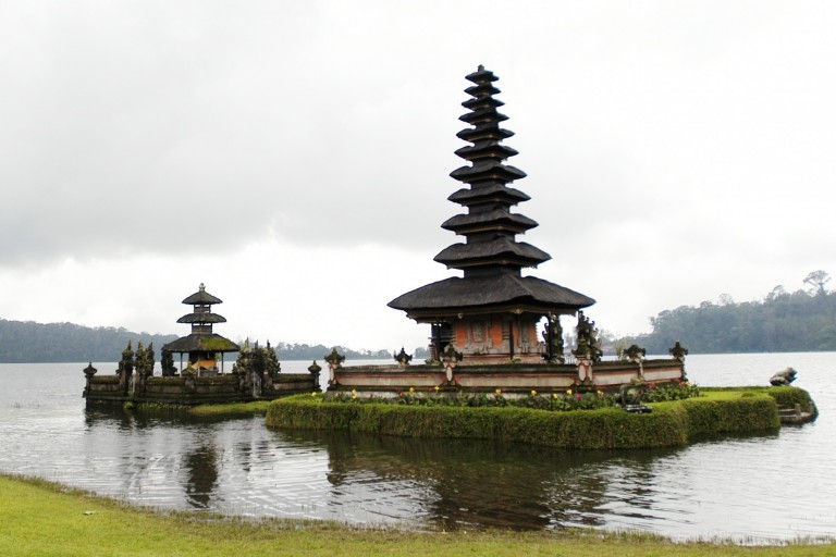 A traditional Balinese temple with multi-tiered roofs stands on a small island in a lake, surrounded by misty hills and cloudy skies. The water reflects the temple’s structure and lush greenery.