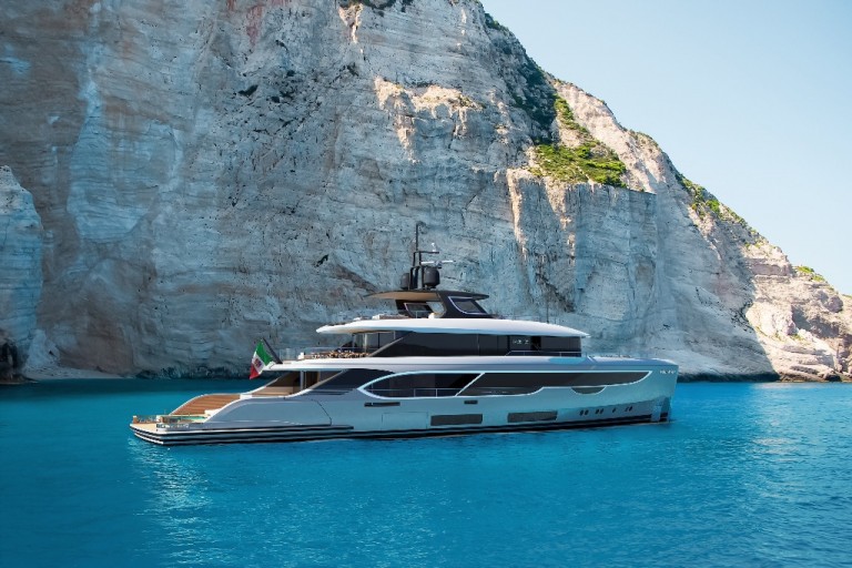 A modern luxury yacht floats on clear turquoise water near tall, sunlit limestone cliffs with patches of greenery under a clear blue sky.