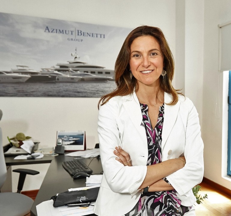 A woman in a white blazer stands smiling with arms crossed in an office, with a desk and computer behind her. A large poster of yachts labelled Azimut Benetti Group hangs on the wall.