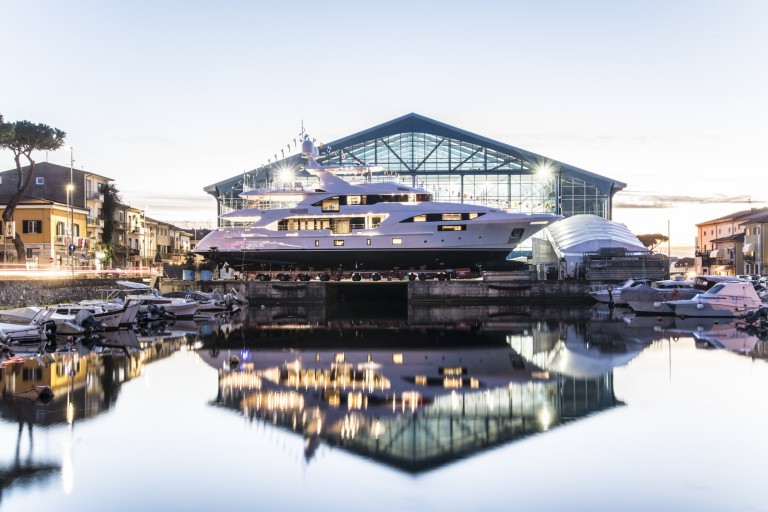A large, white luxury yacht sits moored in front of a modern glass-walled building at sunset, with smaller boats and houses on either side and a clear reflection in the calm water.