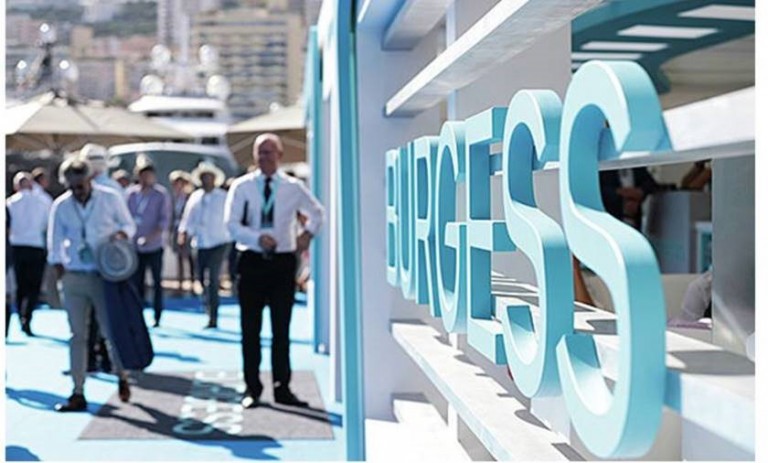 People walk along a blue path at an outdoor event near water, with a large turquoise BURGESS sign prominently displayed in the foreground. The background includes boats and shaded areas.