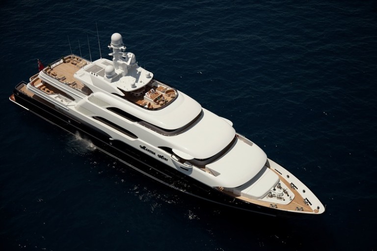 A large luxury yacht with multiple decks sails on calm, dark blue water, viewed from above. The yacht features spacious outdoor seating areas and sleek, modern design.