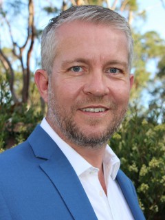 A man with short grey hair and a beard, wearing a blue suit jacket and white shirt, smiles outdoors with green trees and sunlight in the background.
