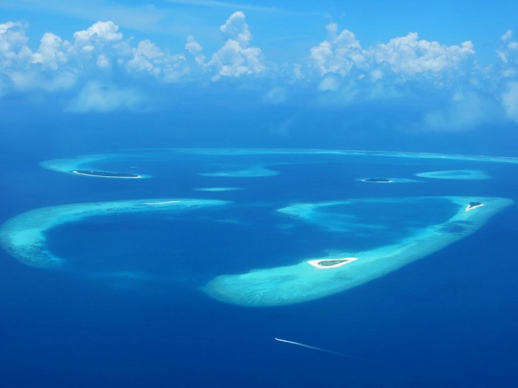 Aerial view of small tropical islands with sandy beaches surrounded by clear blue water; gentle waves and a boat leaving a white trail are visible under a partly cloudy sky.