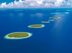 Aerial view of small, round, green islands with sandy shores arranged in a line across deep blue ocean water, under a partly cloudy sky.