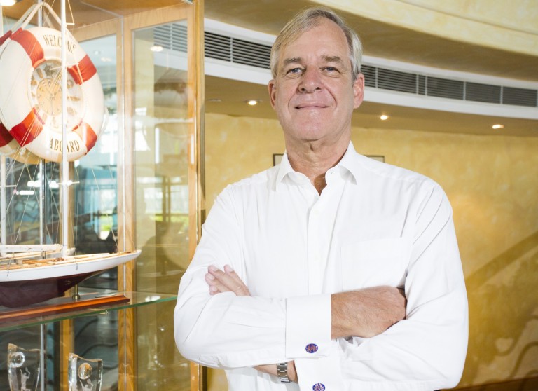 A man in a white dress shirt stands indoors with arms folded, next to a glass display case featuring a model sailing boat and a lifebuoy in the background.