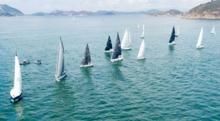 A group of sailboats with white and dark sails participate in a regatta on calm blue water, surrounded by distant hills under a partly cloudy sky.