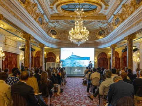A group of people seated in an ornate, chandelier-lit hall watch a presentation about a luxury yacht projected on a large screen at the front of the room.