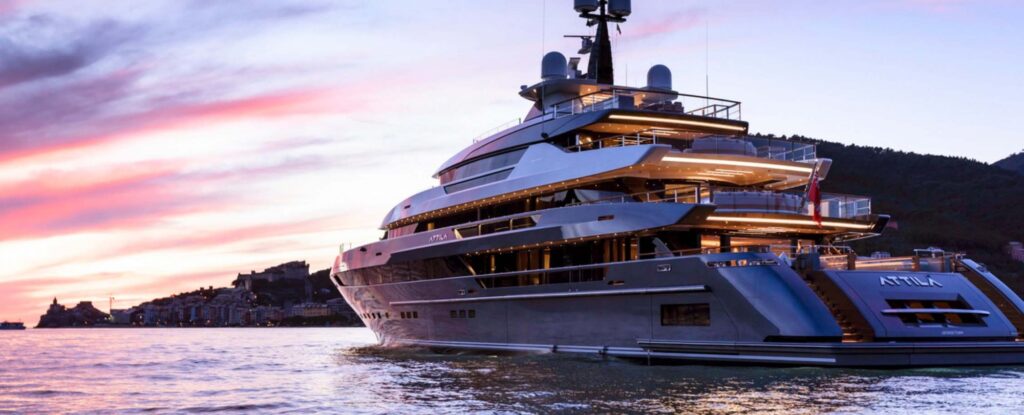 A large, modern yacht named Attila is illuminated and moored on calm water at sunset, with pink and purple skies and distant hills in the background.