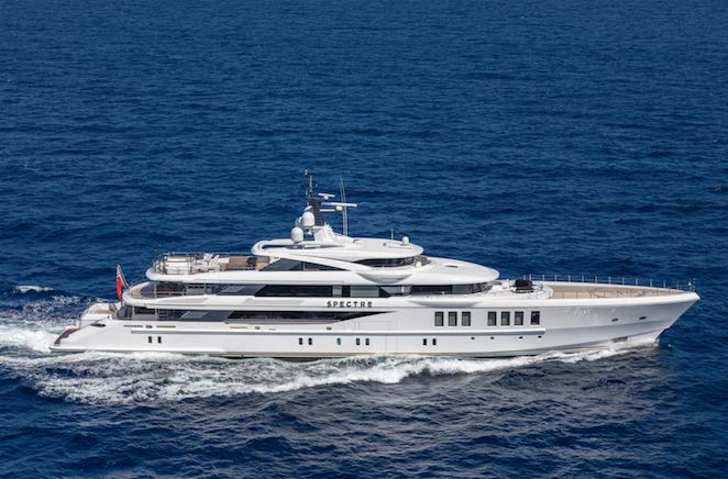 A large white luxury yacht named Spectre sails across a deep blue ocean, leaving a wake behind it, with clear skies reflected on the water.