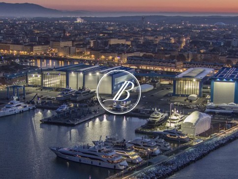 Aerial view of a marina at dusk with several yachts moored near large, illuminated buildings; city lights and mountains are visible in the background. A white circular B logo is centred over the image.