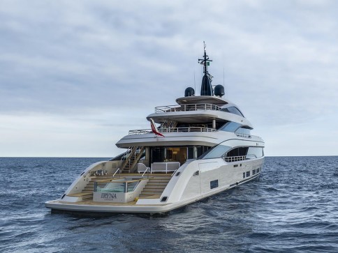 A luxurious white yacht named Irisha is moored on calm water under a cloudy sky, viewed from the stern, showcasing multiple decks and an elegant design.