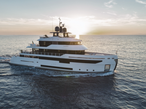 A modern white yacht cruises on calm blue water at sunset, with the sun low on the horizon and soft clouds in the sky, creating a serene and luxurious scene.