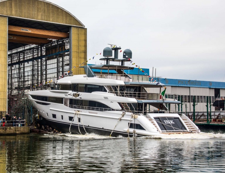 A large white yacht decorated with flags is being launched from a shipyard building into the water, watched by people standing nearby. The scene shows an industrial setting with shipyard structures in the background.