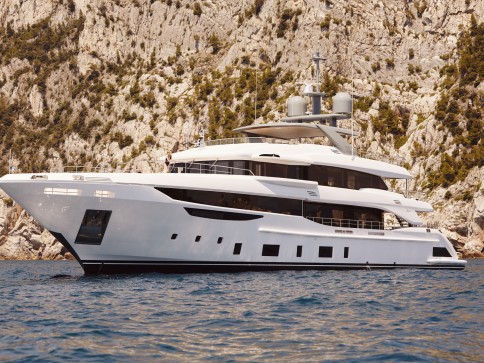 A large, modern white yacht is anchored on calm blue water near a rocky, tree-covered shoreline. The yacht has multiple decks and sleek, contemporary design features.