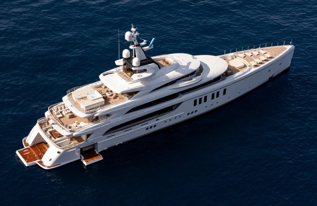 A large, luxurious white yacht is anchored in calm, deep blue water, featuring multiple decks, sun loungers, a pool area, and an open swim platform at the stern.