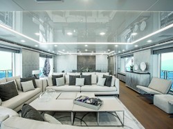 A luxurious yacht lounge featuring modern white sofas with grey and black cushions, a sleek coffee table, large windows with ocean views, stylish decor, and bright recessed lighting.