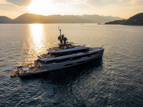 A luxury yacht is anchored on calm water during sunset, with golden sunlight reflecting off the sea and mountains silhouetted in the background.