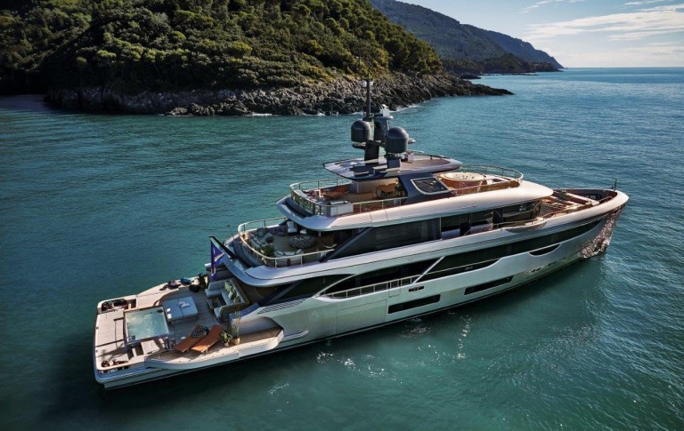 A large, modern luxury yacht with multiple decks floats on calm turquoise water near a rocky, forested shoreline under clear skies.