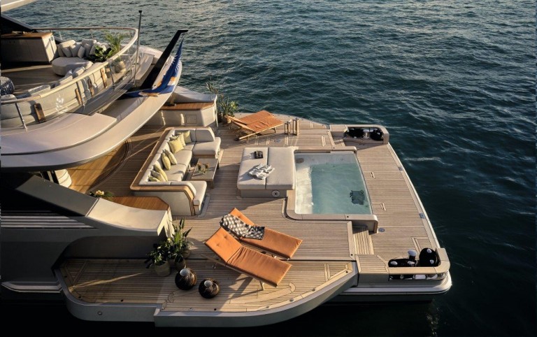 Luxury yacht deck with lounge seating, a hot tub, two orange sun loungers, plants, and decor, overlooking calm blue water in the evening light.