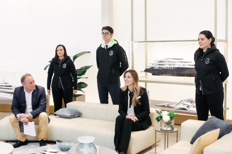 Five people are gathered in a modern, bright room with white walls and sofas. Three stand in matching black attire, while two sit. A model yacht and books are displayed behind them. The atmosphere appears professional.