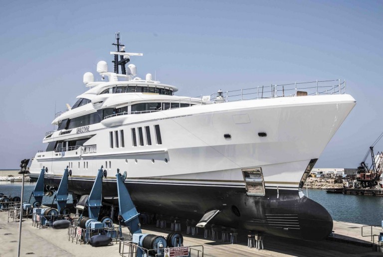 A large, white luxury yacht is lifted out of the water on mechanical supports at a shipyard, with a clear sky and some cranes and construction materials in the background.