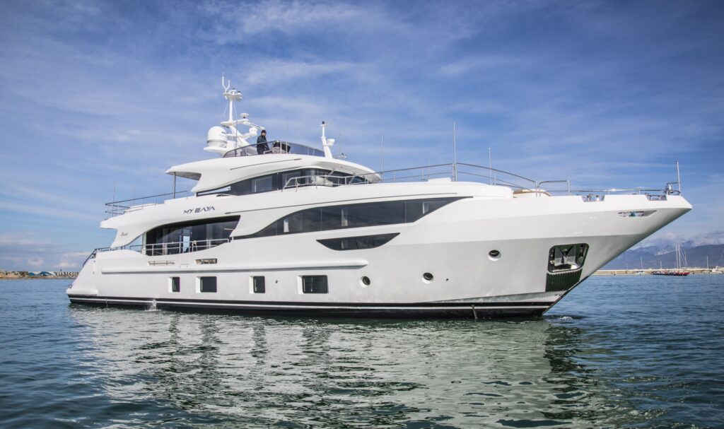 A large, modern white yacht floats on calm water under a blue sky with light clouds, with distant land and mountains visible in the background.