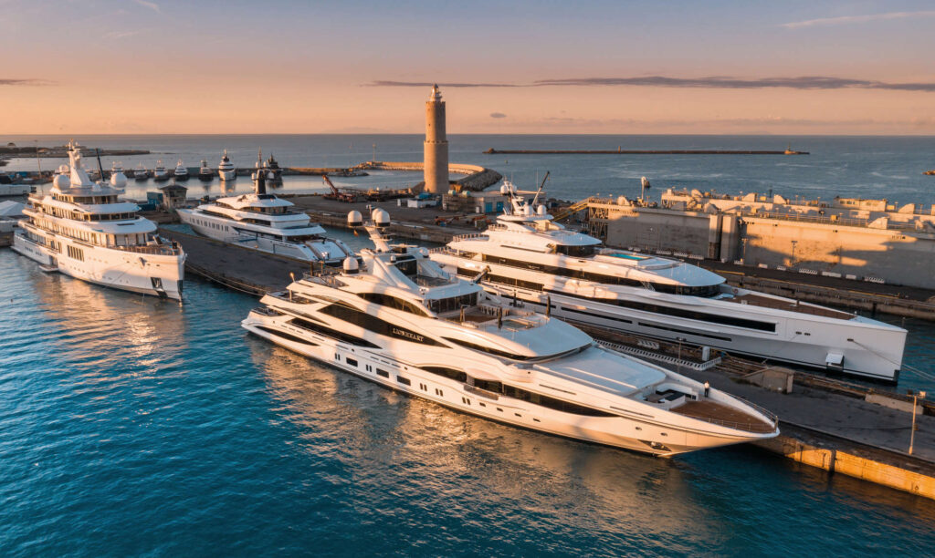 Several large luxury yachts are moored at a marina during sunset, with calm blue water and a lighthouse in the background. The scene has a warm, golden glow from the setting sun.