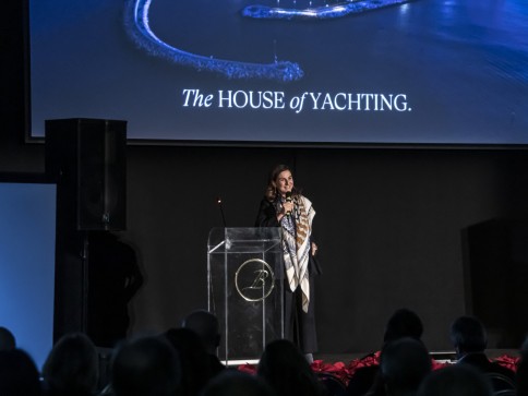 A woman stands at a clear lectern on stage, speaking to an audience. Behind her is a large screen displaying an aerial view of a marina and the text The HOUSE of YACHTING.