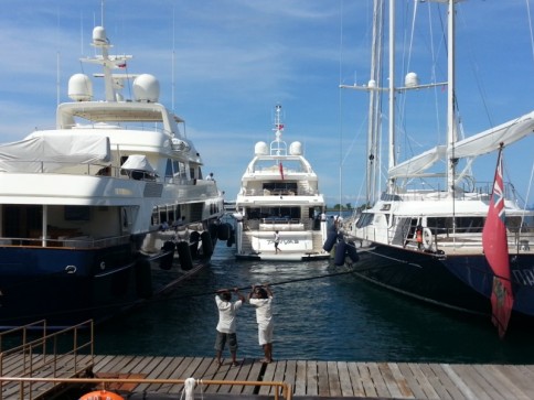 Two people in white uniforms are pulling a heavy rope on a quay between large yachts moored in a sunny marina. The water is calm, and the sky is clear with a few clouds.