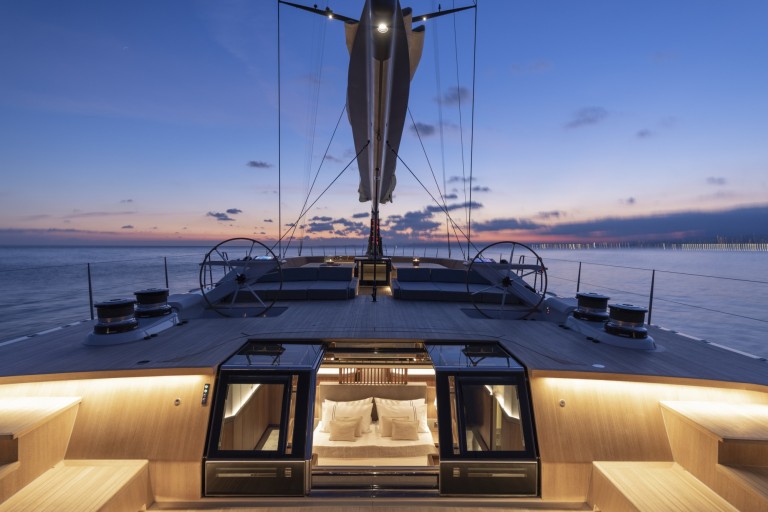 A luxurious yacht deck at sunset with glowing lights, dual steering wheels, and a sail above. Below deck, a cosy, well-lit bedroom with a double bed is visible through open glass doors. Calm sea and colourful sky in the background.