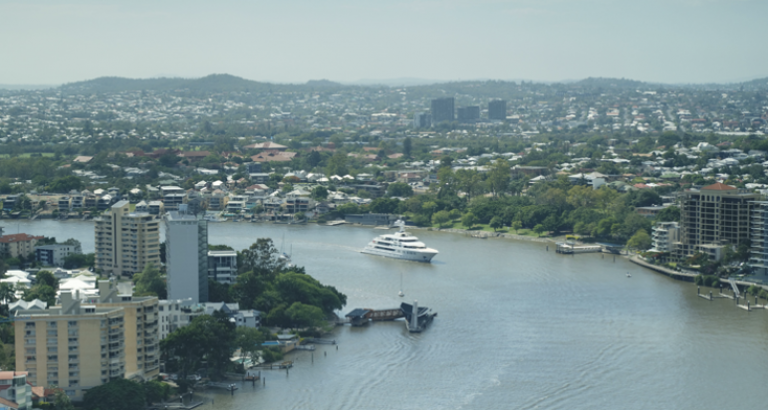 A large white yacht sails along a winding river bordered by city buildings and green trees, with a sprawling urban landscape and hills visible in the background.