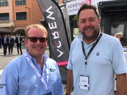 Two men wearing conference badges and sunglasses smile outdoors at a business event. One wears a light blue shirt; the other a light blue polo shirt. A business flag and several people are visible in the background.