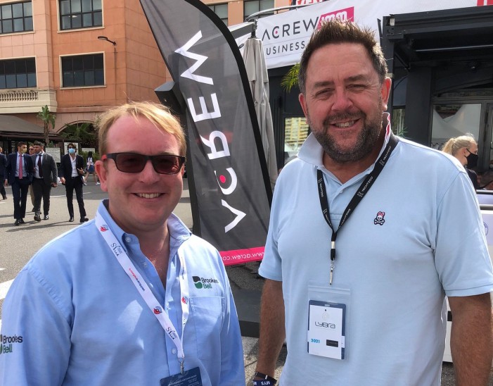 Two men wearing sunglasses and lanyards stand smiling outdoors at an event, with branded flags and other attendees visible in the background.