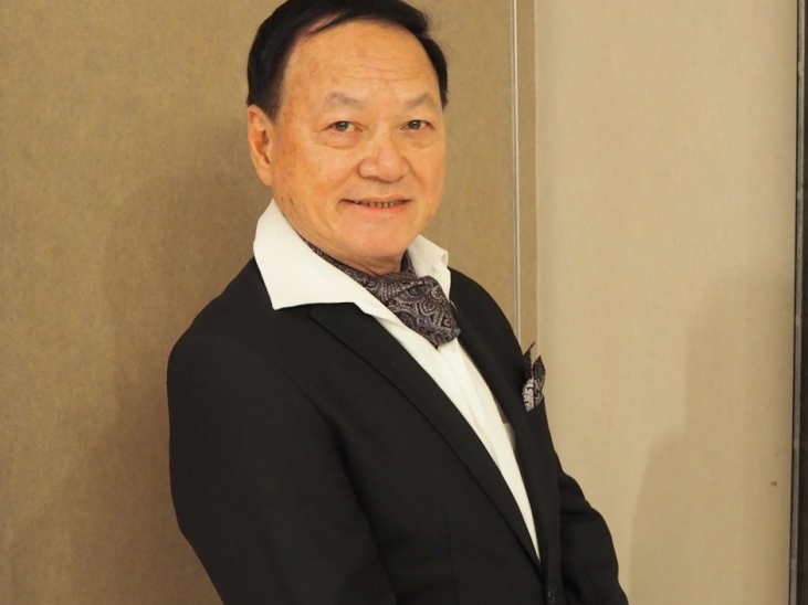 A man in a black suit with a white shirt and patterned scarf smiles whilst standing against a beige wall.