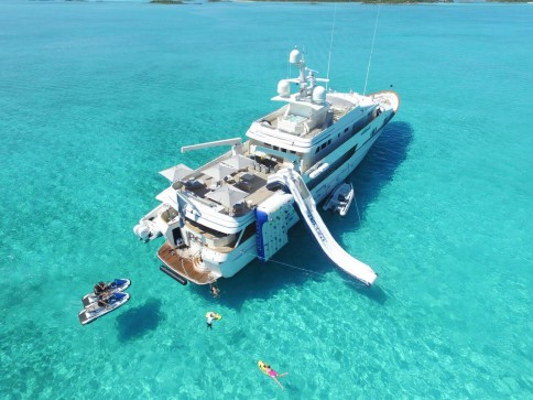 A large white yacht is anchored in clear turquoise water. People are swimming, kayaking, and riding a jet ski nearby. The yacht has a slide and a climbing wall extending into the water. Islands are visible in the distance.