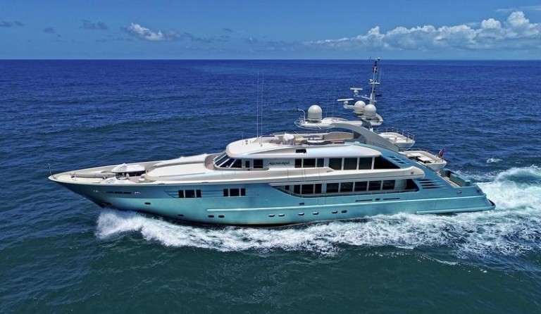 A large, modern luxury yacht sails on the open sea under a clear blue sky, creating white waves as it moves swiftly through the water.