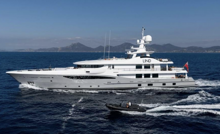 A large white luxury yacht named LIND sails on calm blue water with a small motorboat nearby. Green mountains and clear blue sky are visible in the background.